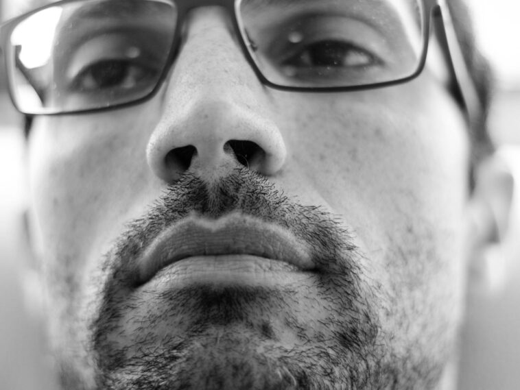 Black and white close-up photo of a bearded man wearing glasses, looking thoughtful.