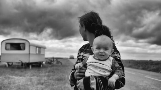gray scale photography of woman carrying baby looking at camper trailer